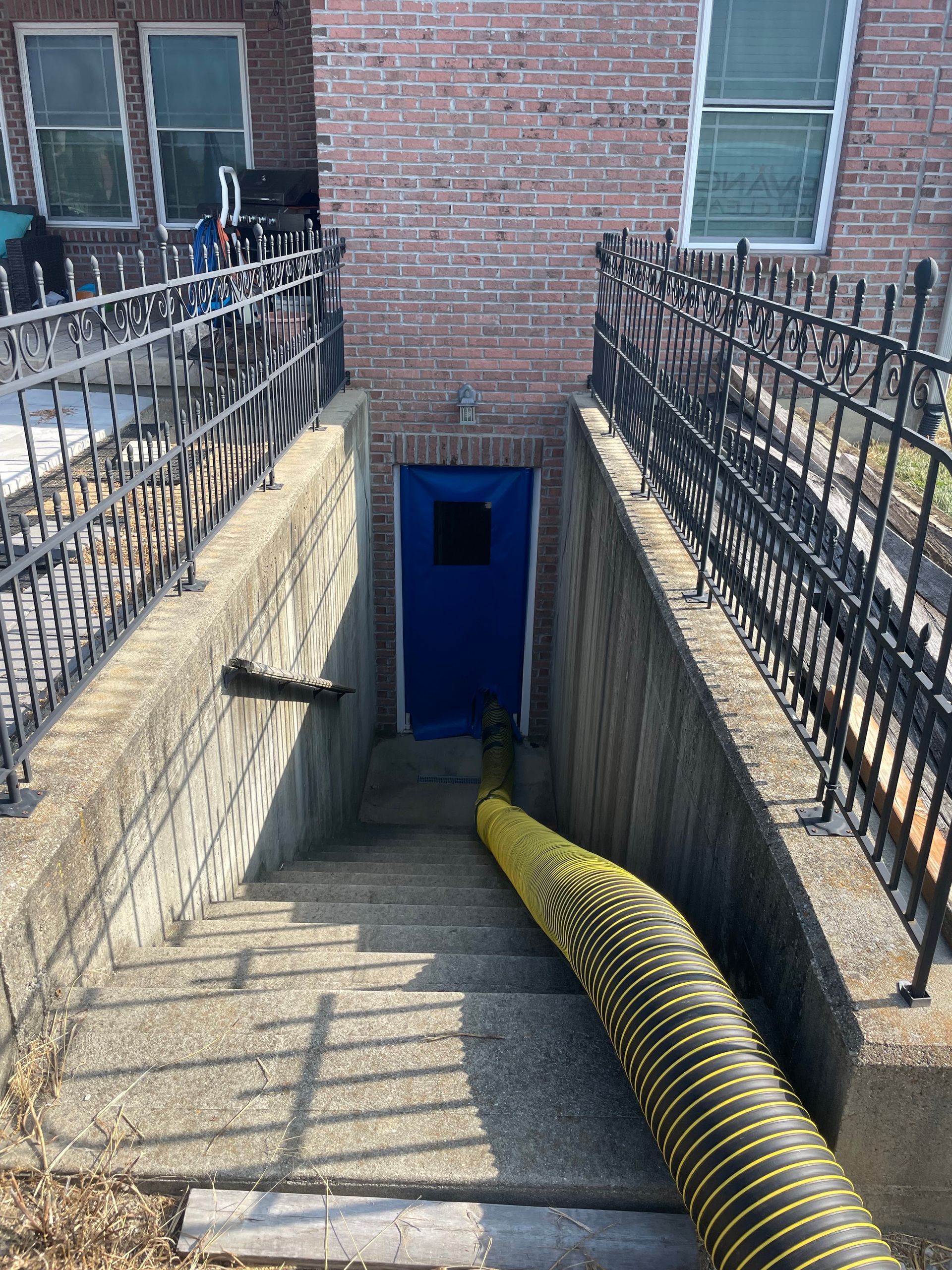 Stairwell with black railings leading down to a blue door beside a brick building, with a yellow hose on the steps