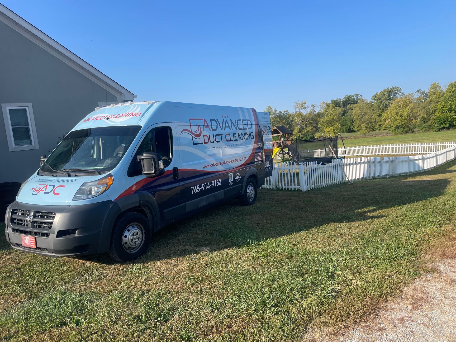 Blue rental van parked beside a building and white fence on a sunny grassy lot