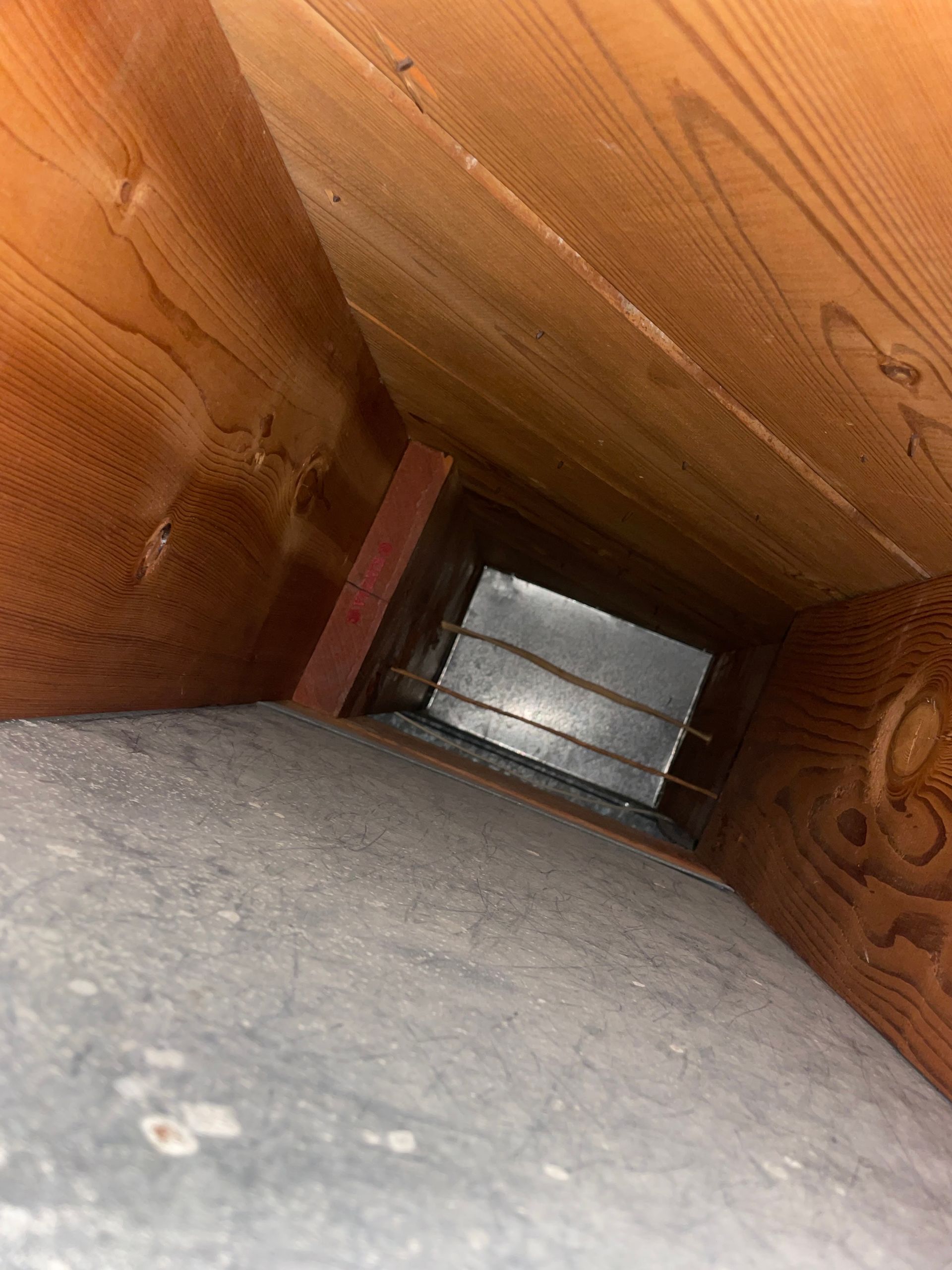 View down a narrow wooden vent or duct toward a small metal opening at the end