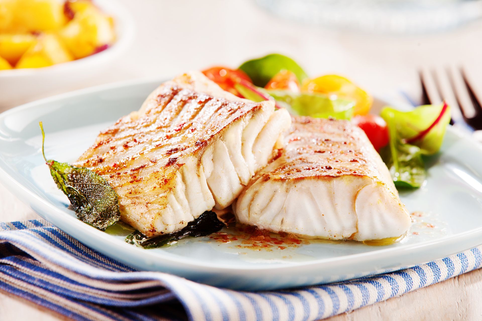 Grilled fish fillets on a white plate with asparagus and salad on a striped cloth