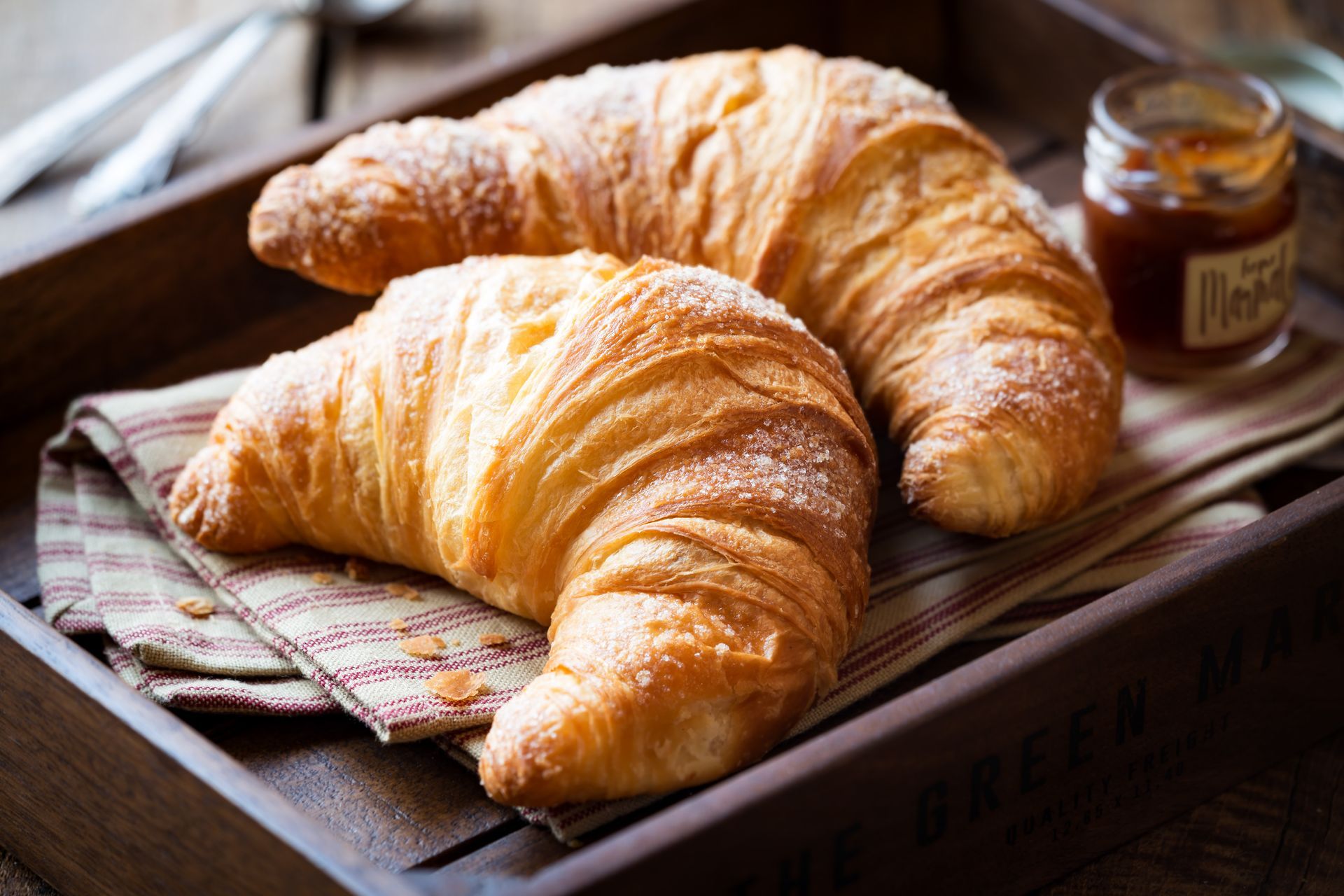 Two golden-brown croissants on a tray with a striped cloth and a jar of jam nearby