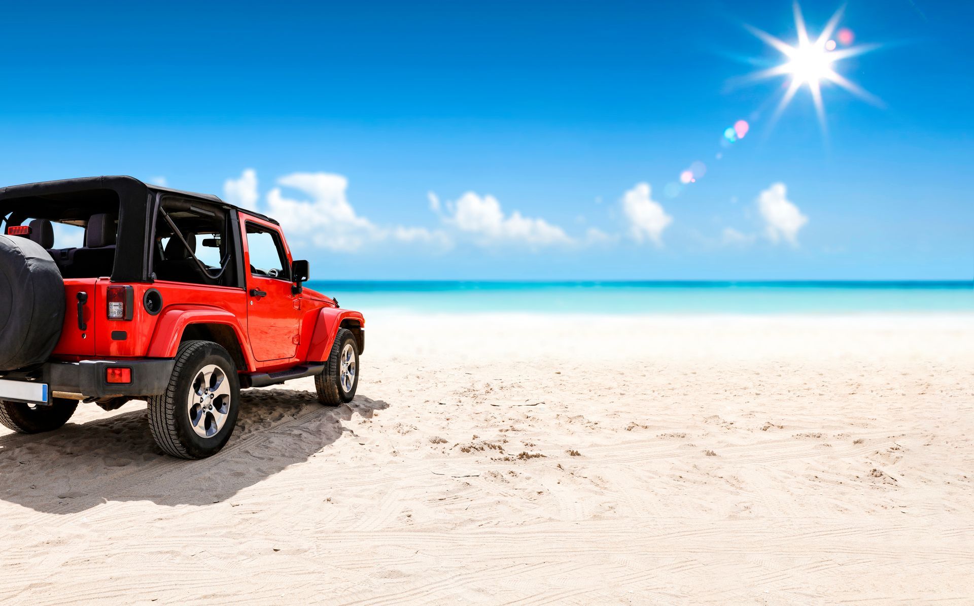 Red Jeep parked on a bright sandy beach beside turquoise water under a sunny blue sky