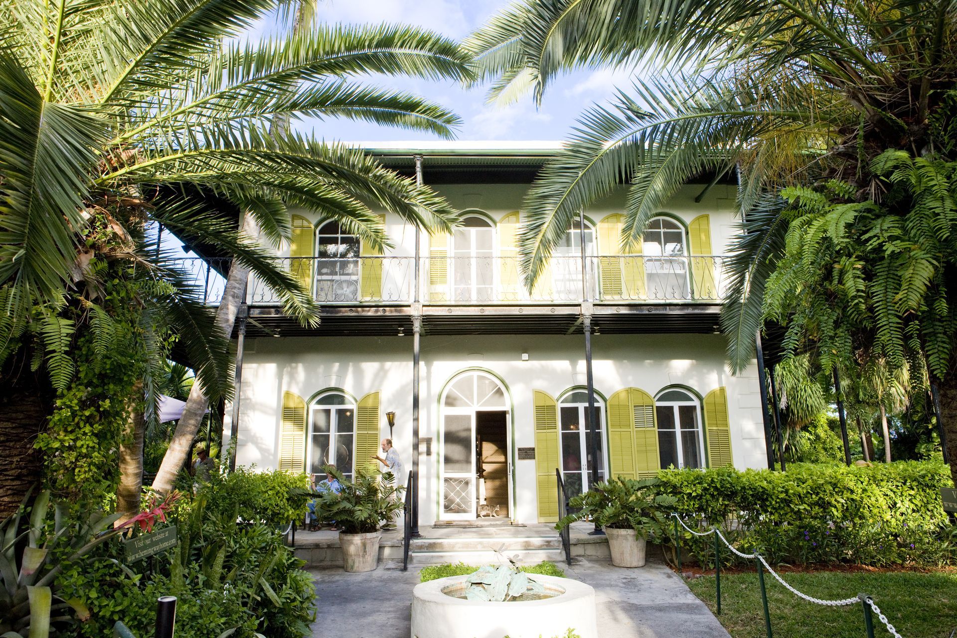 Two-story white house with balcony, framed by palm trees and lush greenery.