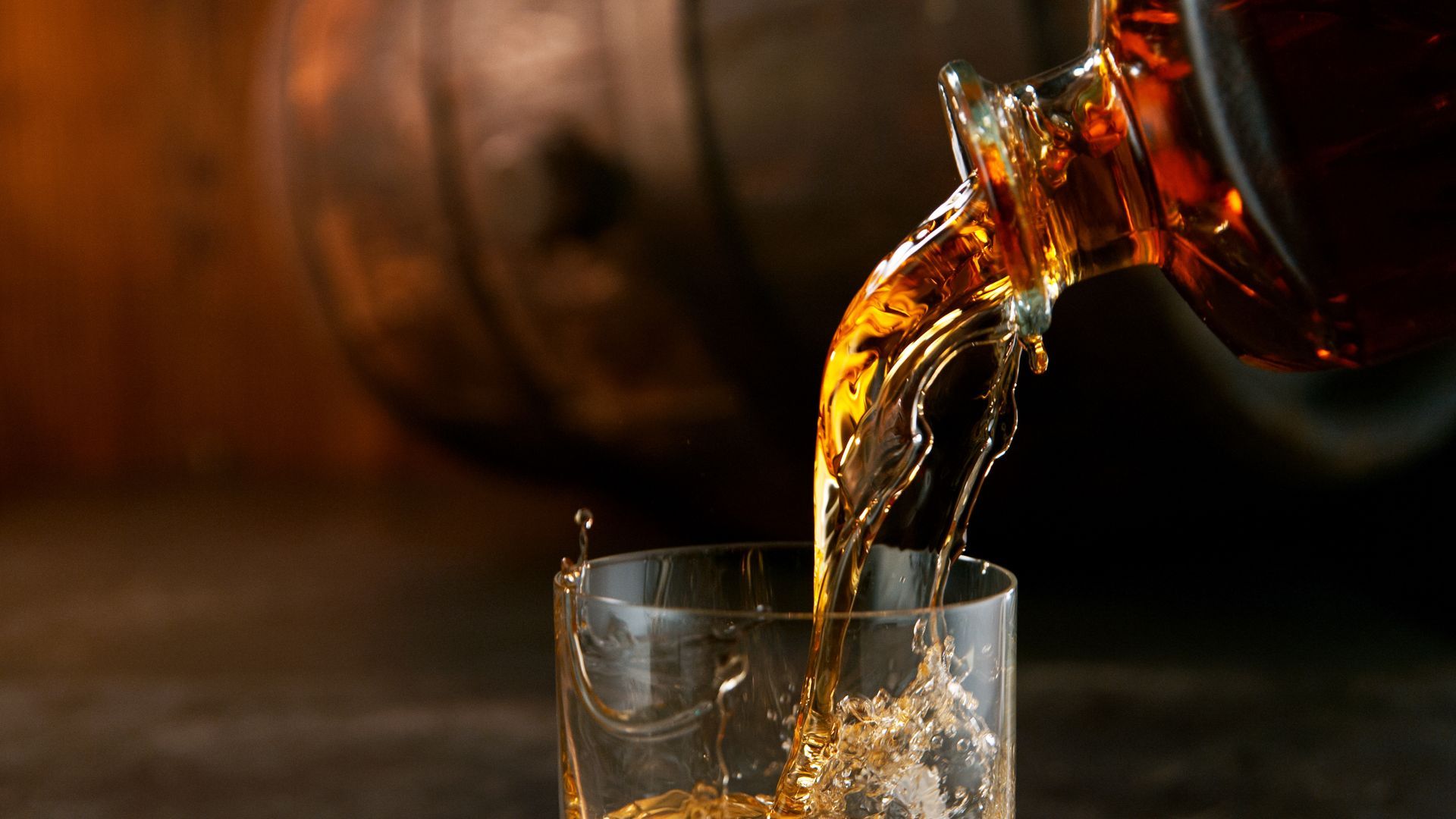 Dark amber liquid pouring into a glass with ice, against a blurred wooden barrel background