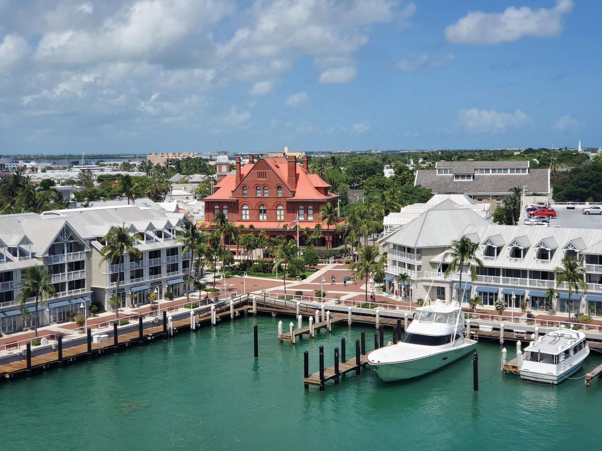Marina with white buildings, red-roofed house, docked boats, and turquoise water under a blue sky