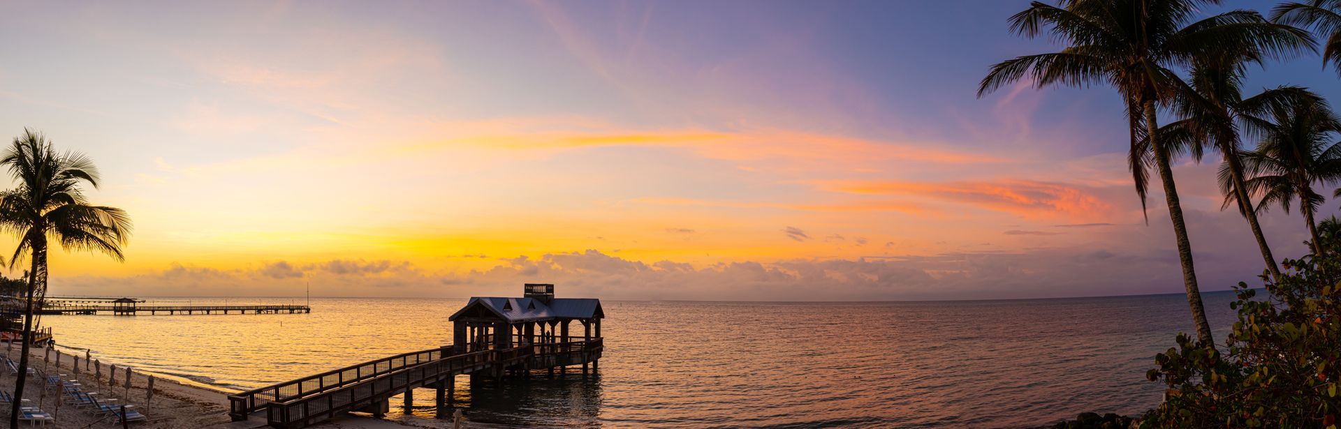 Sunset over a calm ocean with a pier, palm trees silhouetted at the shore, and a small hut on the water