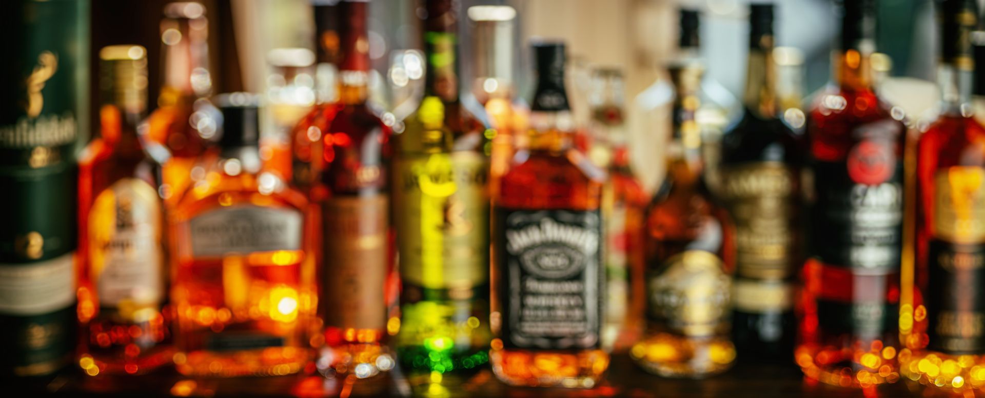 Assorted liquor bottles lined up on a bar shelf, glowing in warm orange light