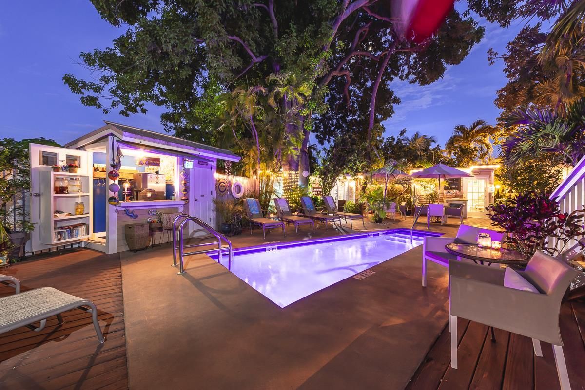 Backyard patio with illuminated pool, lounge chairs, and a lit house at dusk