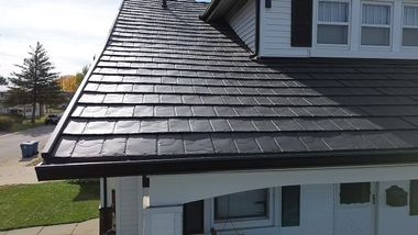 Close-up of a dark slate roof on a white house with a small upper window and trees in the background