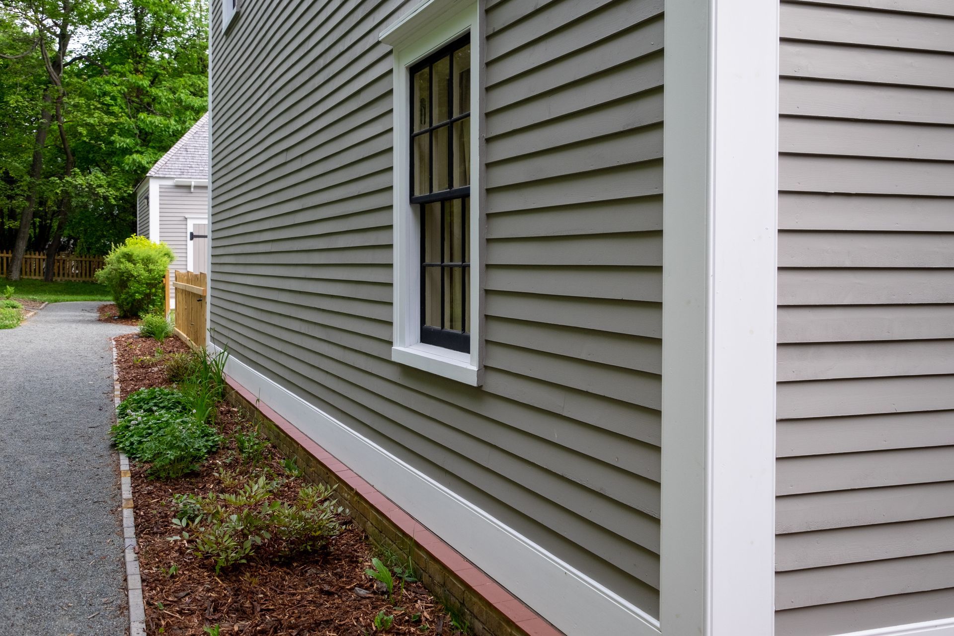 Side of a gray house with white trim, narrow window, gravel path, and landscaped flower beds
