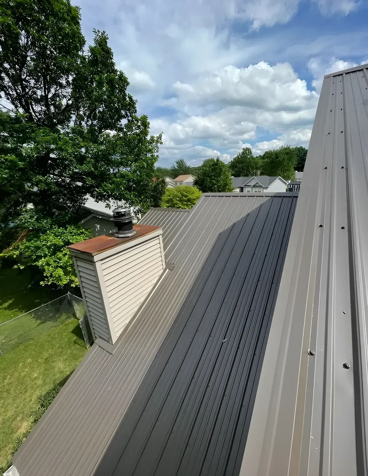 Rooftop with gray metal panels, white chimney, and leafy trees under a partly cloudy sky