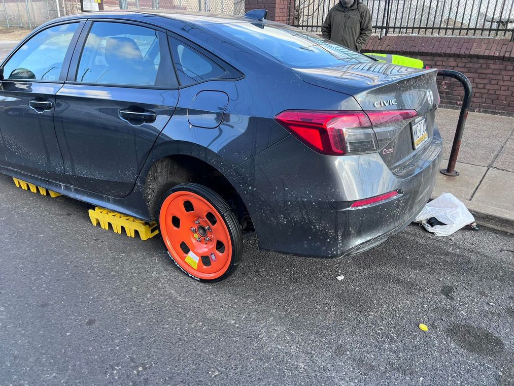 Black sedan with bright orange rear wheel and yellow wheel chocks parked beside a wall