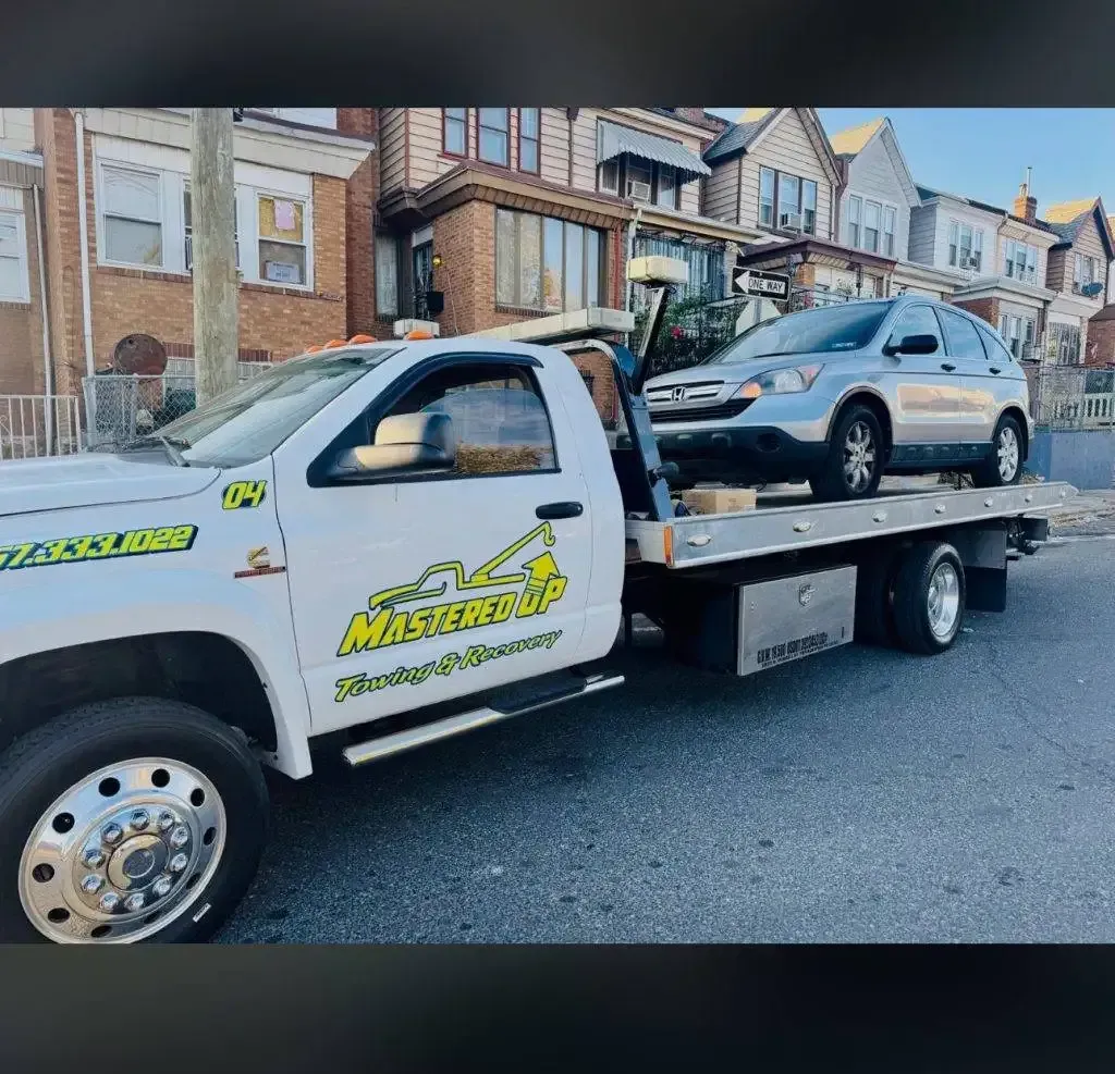 Tow truck carrying a silver car on a residential street lined with brick houses.