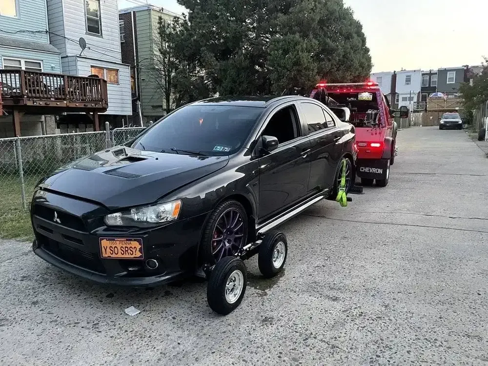 Black sports car towed on a dolly behind a red tow truck on a residential street.