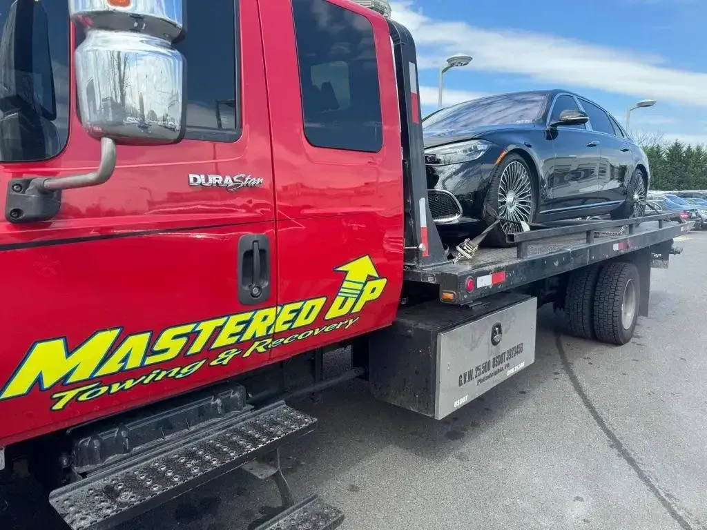 Red tow truck hauling a black car on a flatbed in a parking lot.