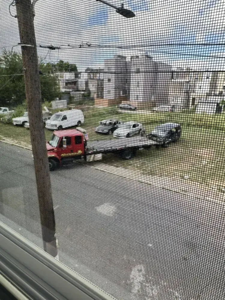 Red tow truck hauling a silver car on a street, viewed through a window screen.