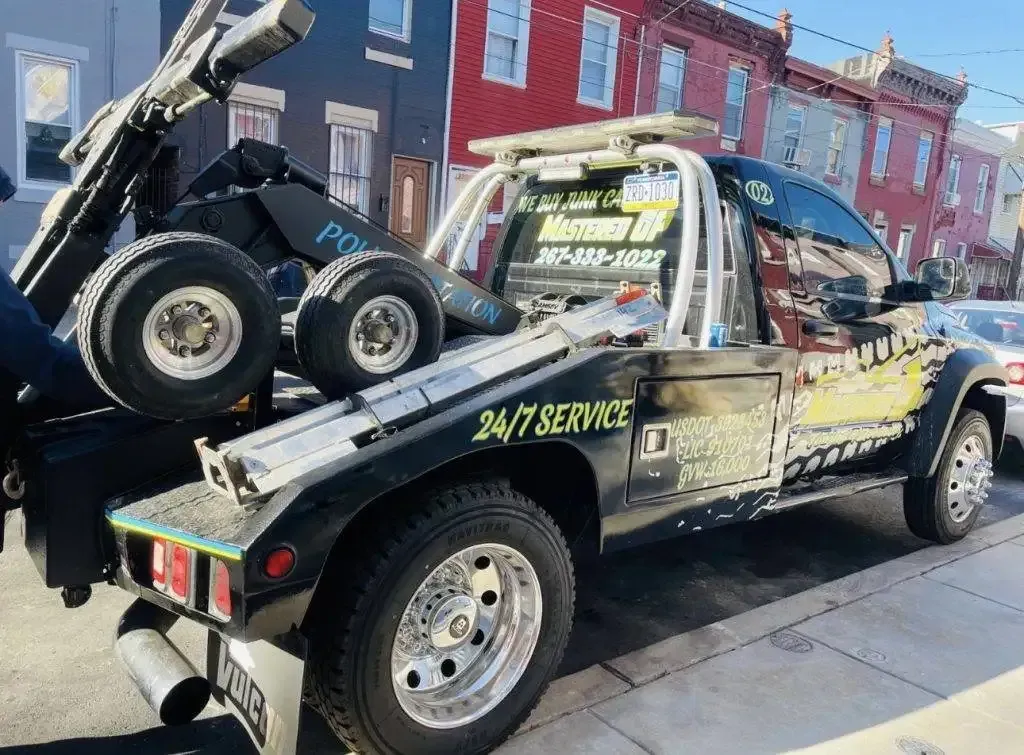 Tow truck hauling a damaged car on a city street, with 24/7 service lettering.