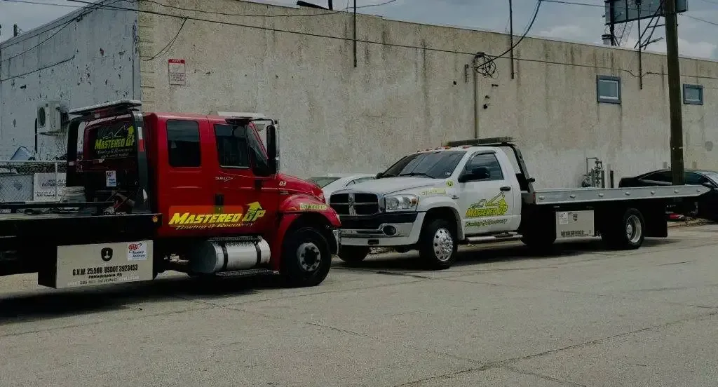 Red tow truck hauling a white flatbed truck on a street beside a concrete wall.
