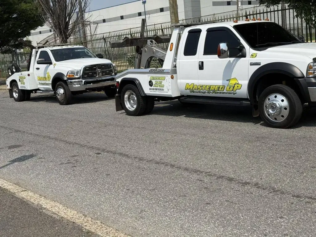 Two white utility trucks parked on a street, one towing the other, beside a building.