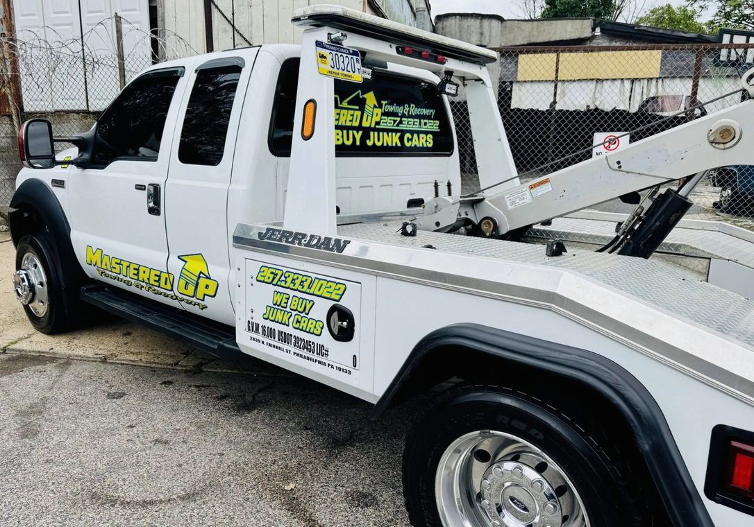 White tow truck with “Buy Junk Cars” signs parked outdoors near a fence.