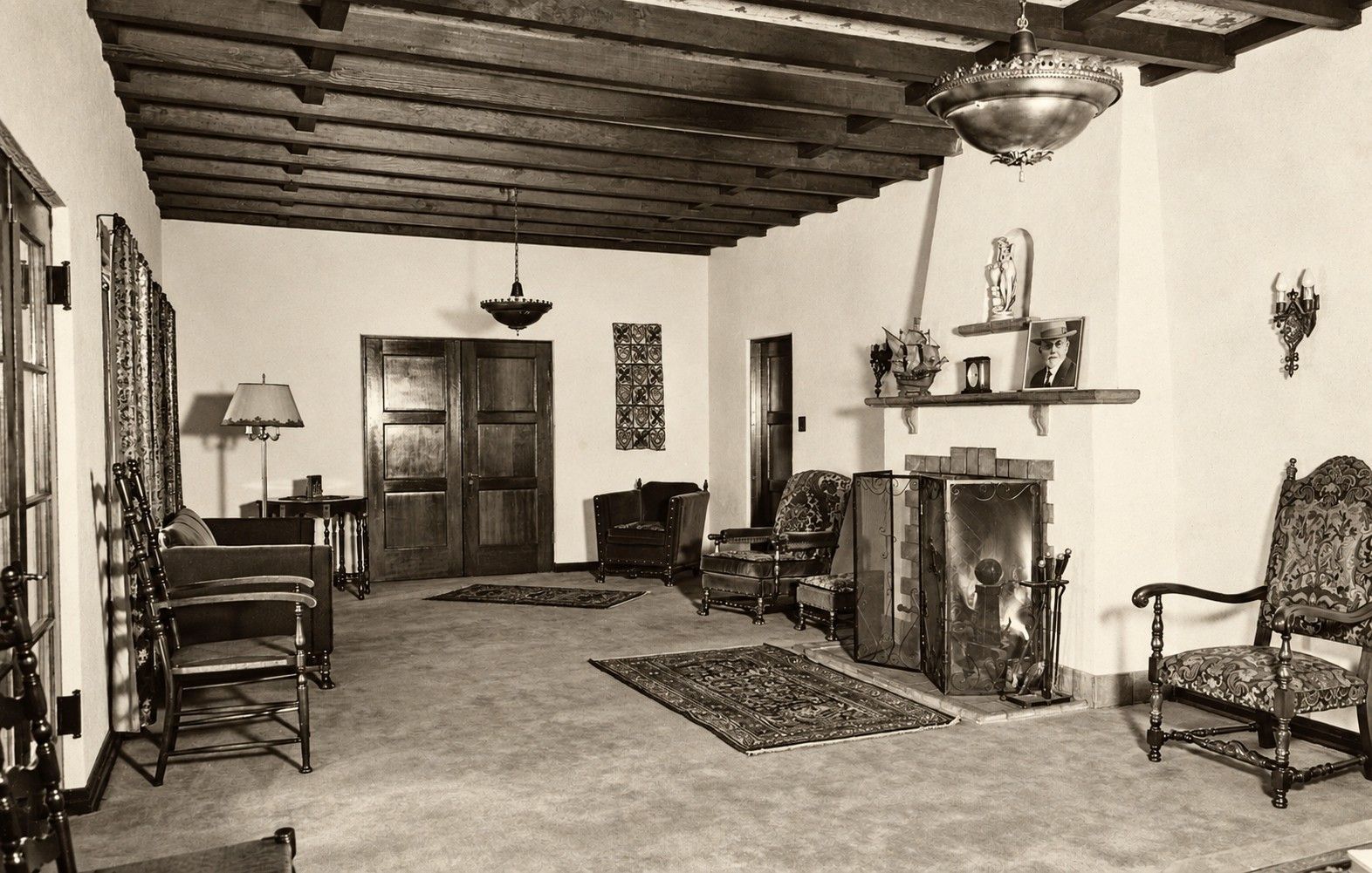 Sepia-toned living room with wooden ceiling beams, fireplace, rocking chairs, and rugs