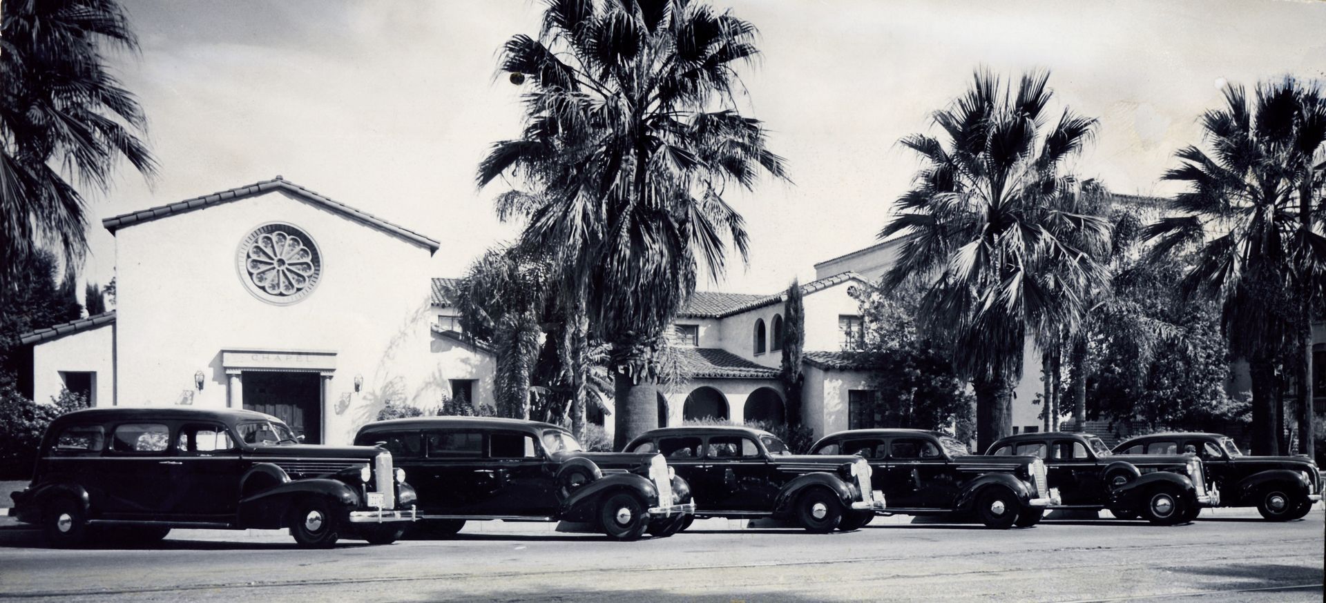 Black-and-white photo of a building with palm trees and a row of parked vintage cars