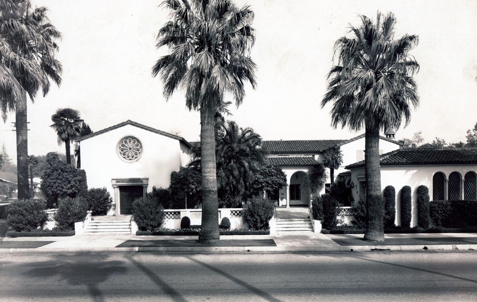Black-and-white view of a palm-lined suburban house with a fountain and arched porch.