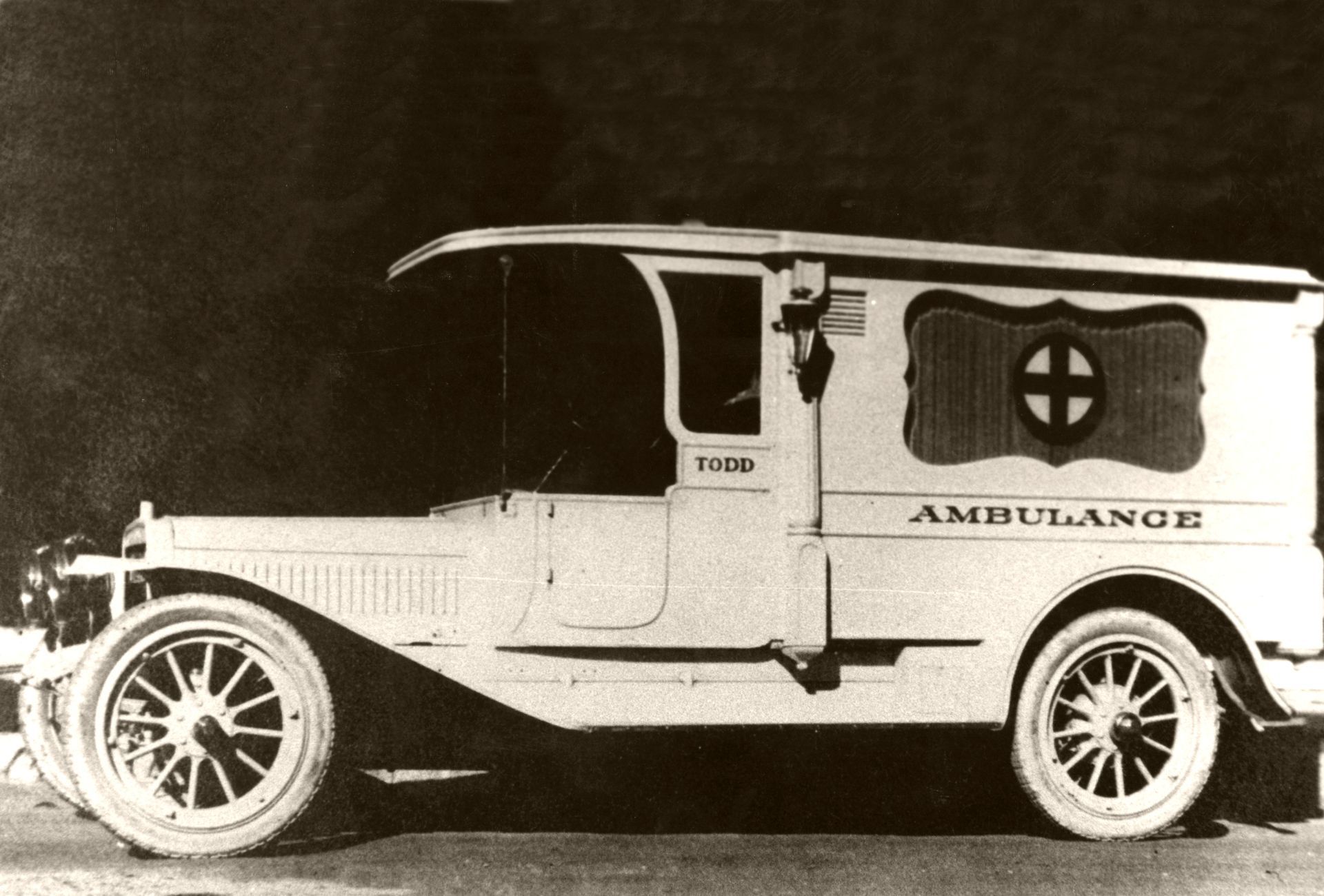 Vintage white ambulance car with boxy body and large spoked wheels, side view on dark background