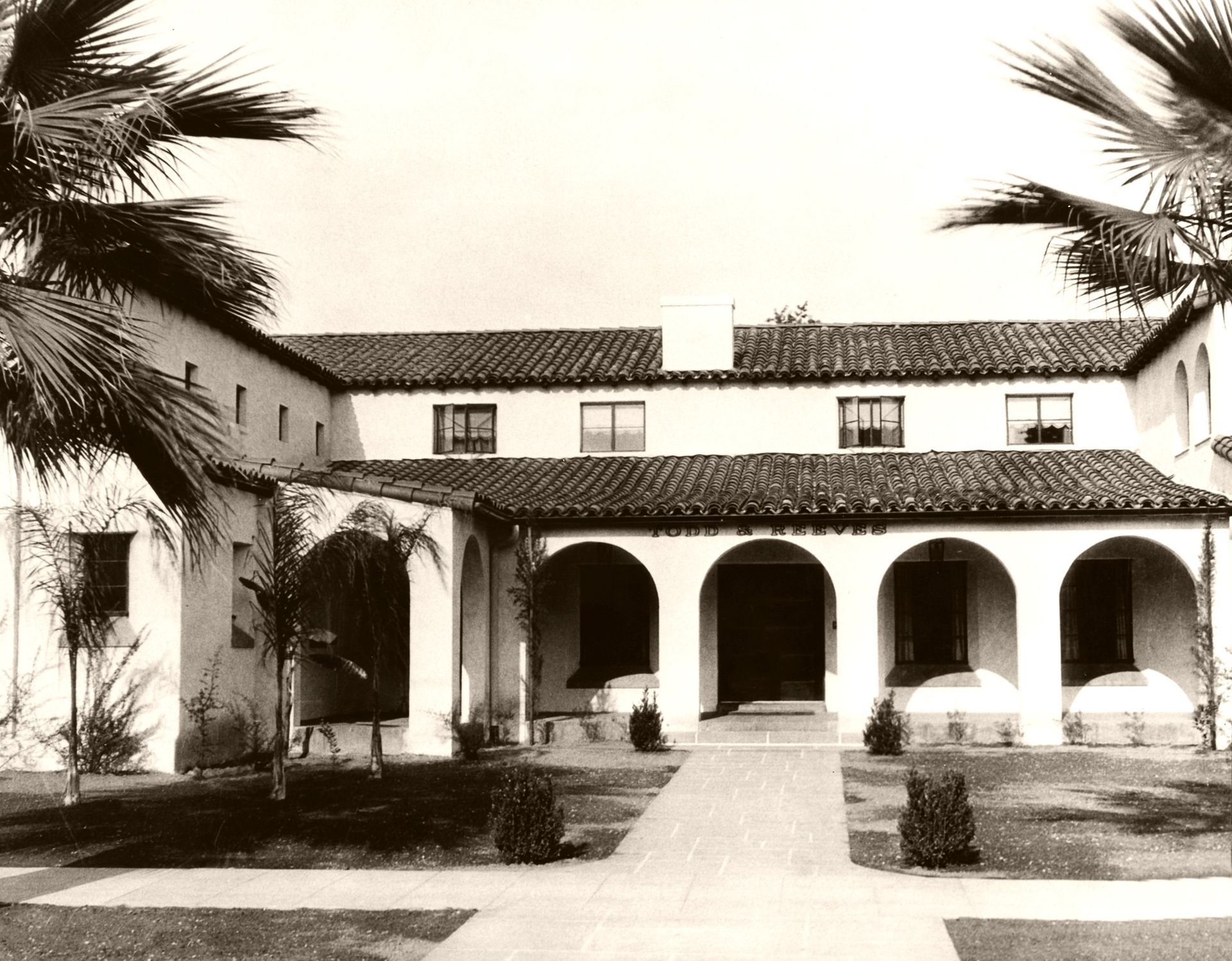 Sepia-toned courtyard with arched colonnade, tiled roof, and palm trees framing a historic building.