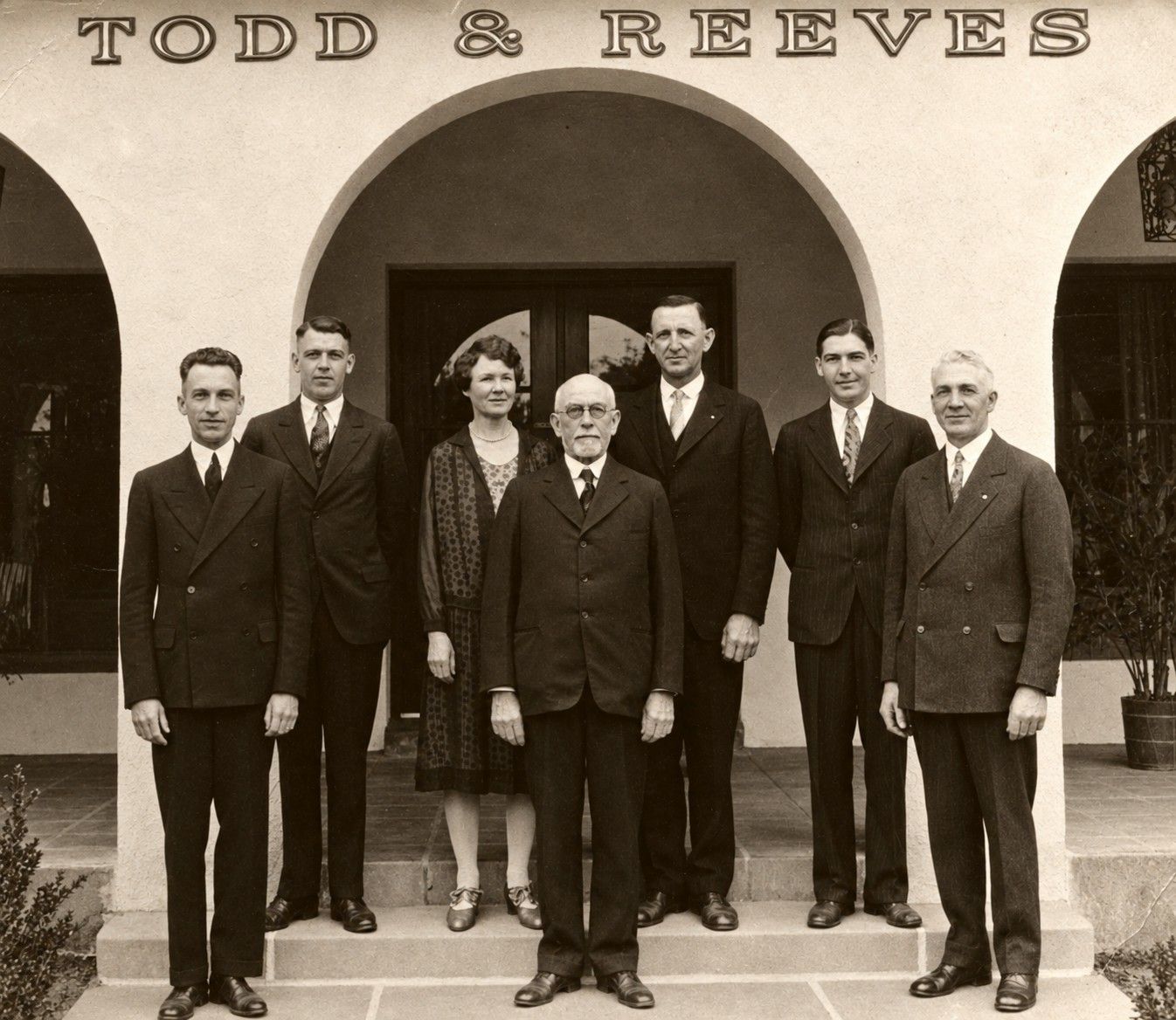 Seven people in suits posing outside the Todd & Reeves storefront under arched columns.
