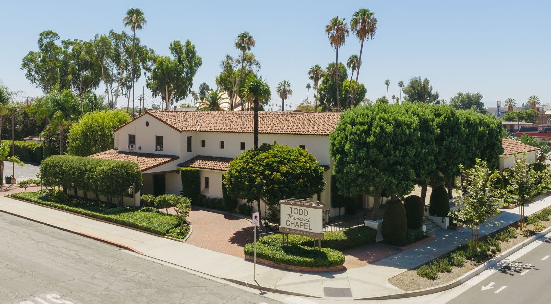 Spanish-style home on a corner lot, surrounded by trees and hedges on a sunny street