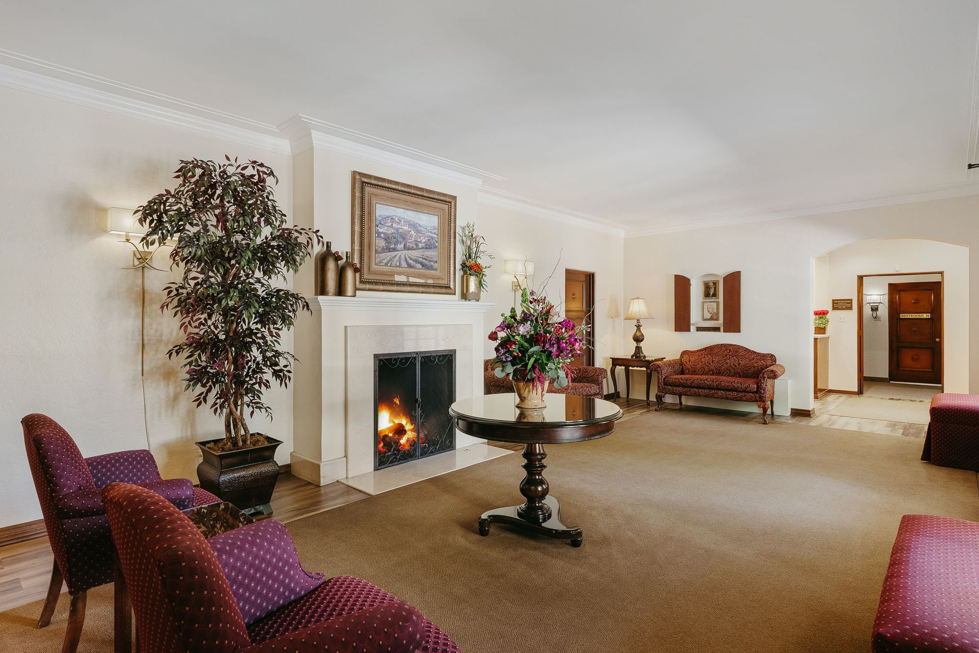 Elegant living room with fireplace, floral arrangement, burgundy chairs, and beige carpet.