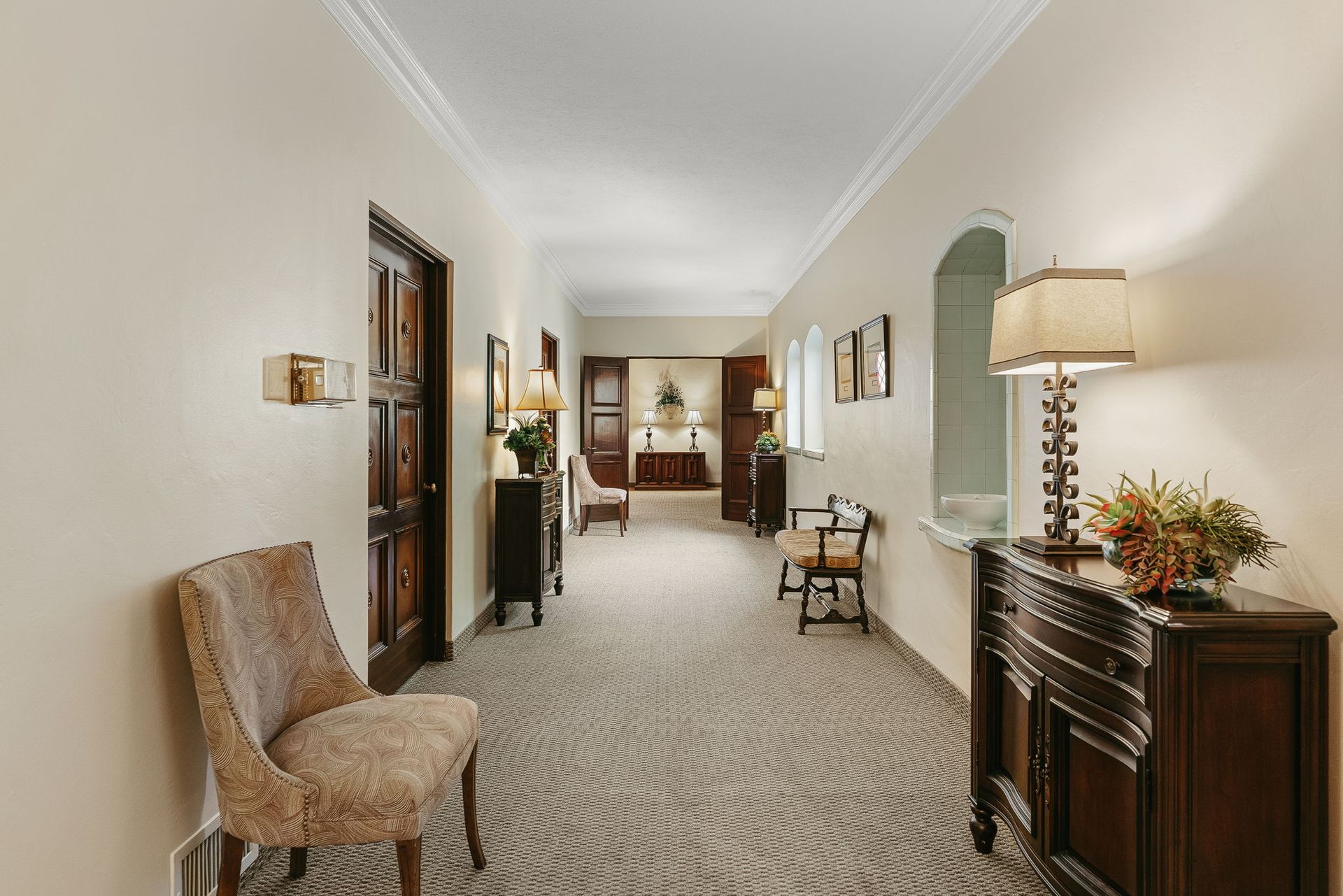 Elegant hallway with patterned carpet, wall sconces, chairs, and a wooden console table with lamp and flowers