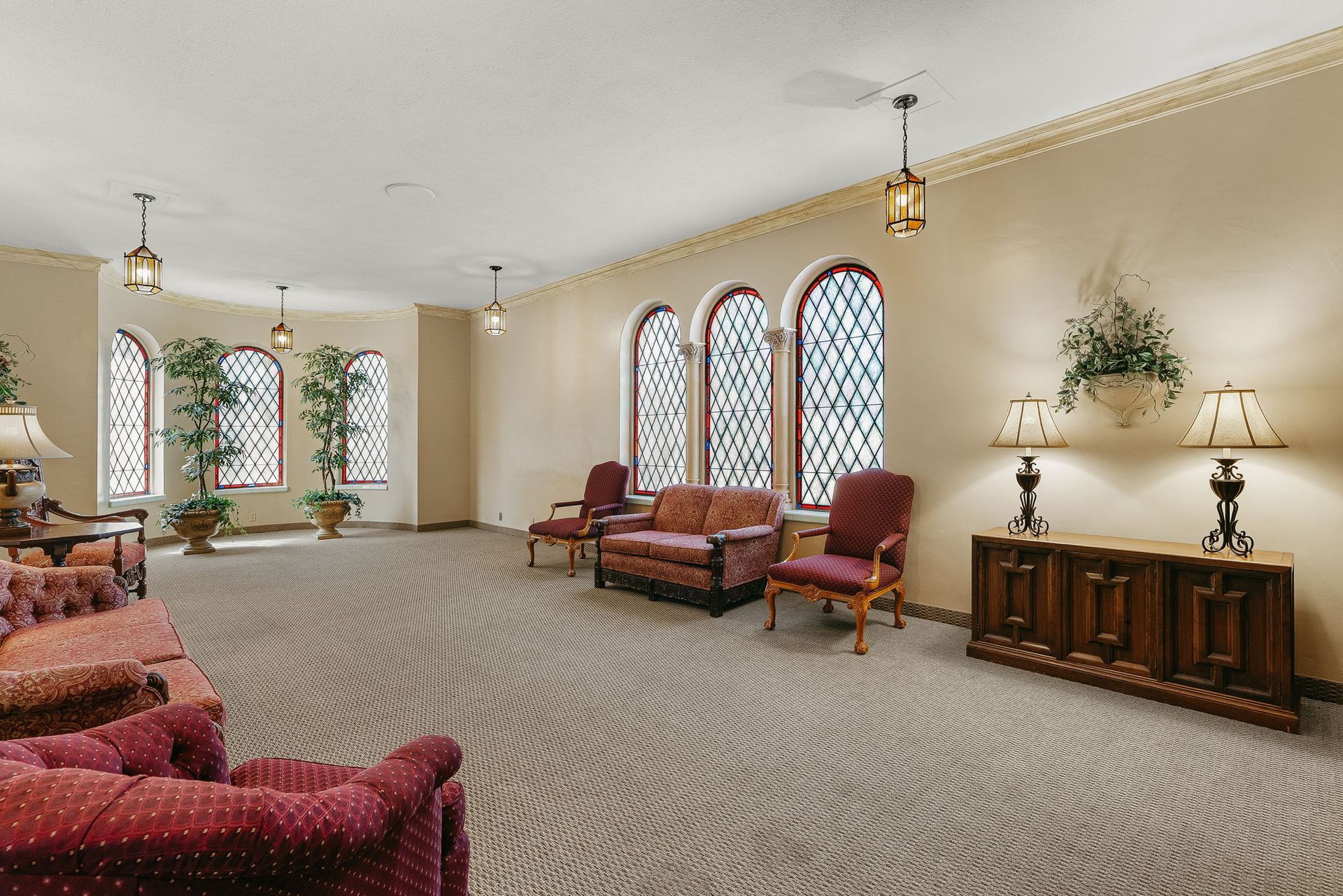 Spacious living room with arched windows, patterned carpet, red chairs, and wooden sideboard.