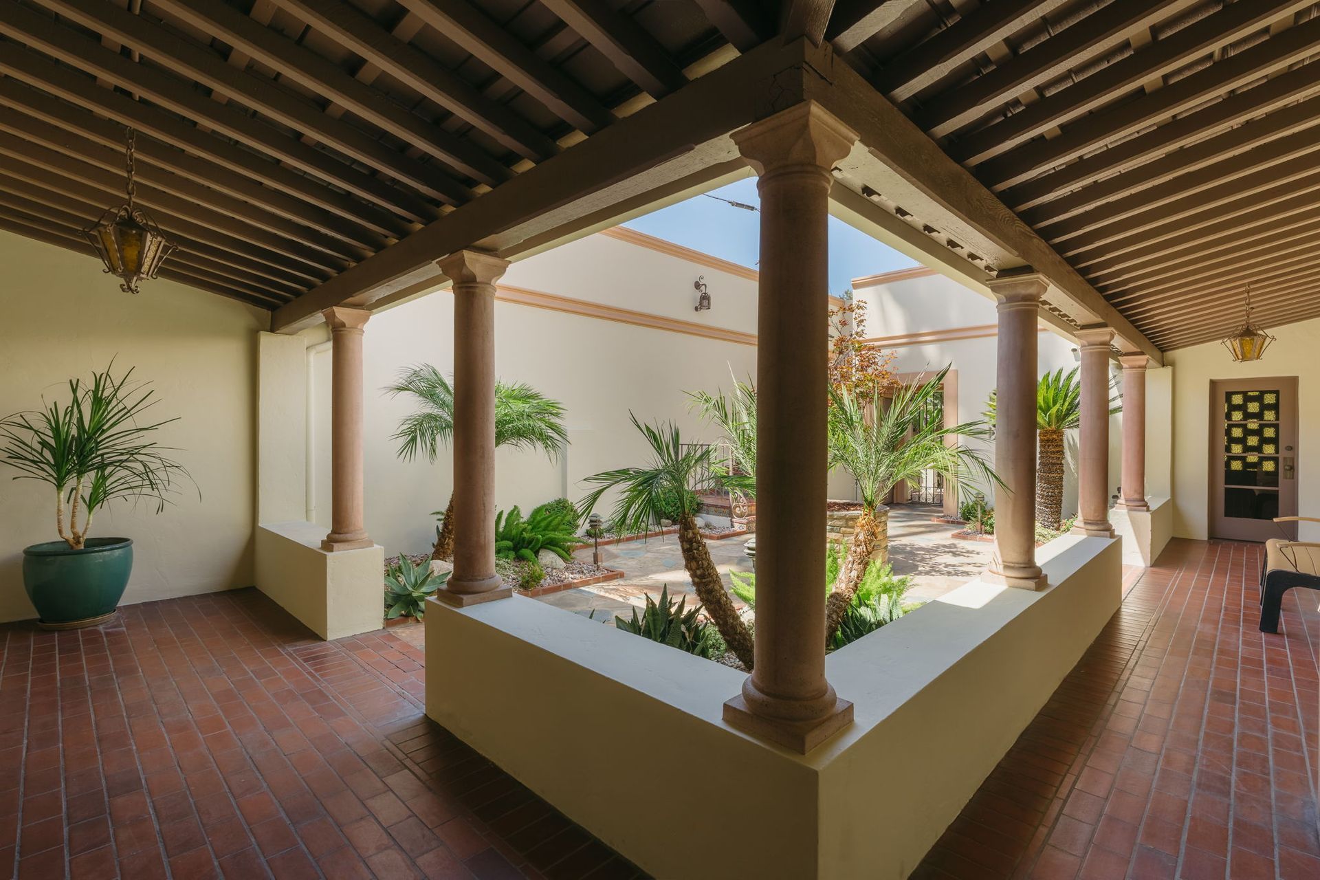 Covered courtyard with brick floor, central planter, and potted tropical plants under wooden beams