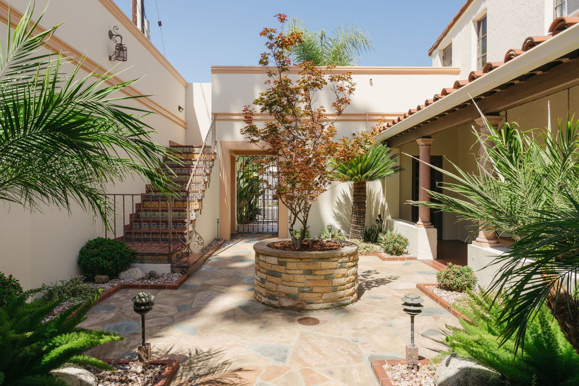 Sunlit courtyard with stone fountain, stairs, potted tree, and desert plants beside white stucco walls