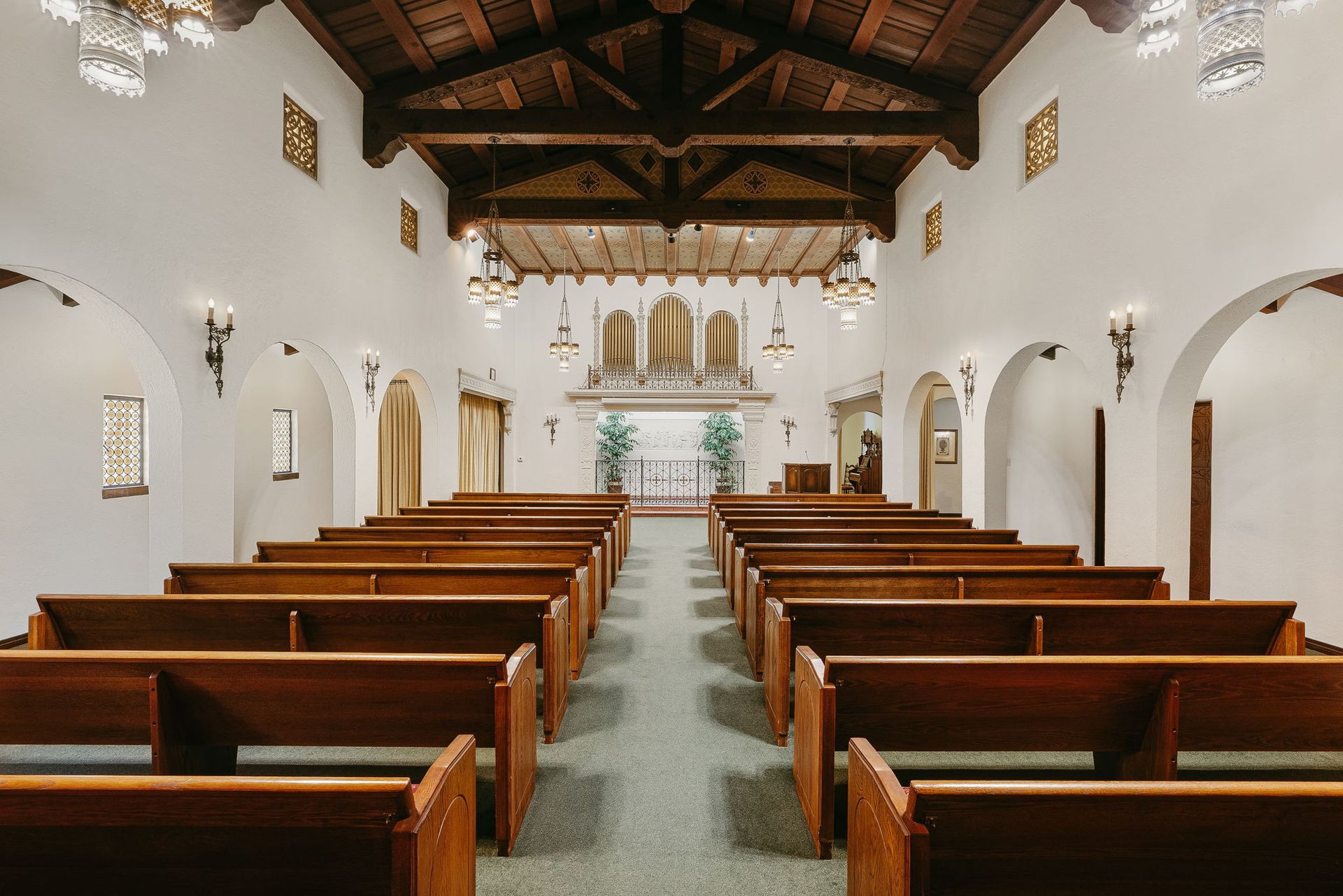 Interior of a church with wooden pews, white walls, and a vaulted wooden ceiling, viewed from the back.