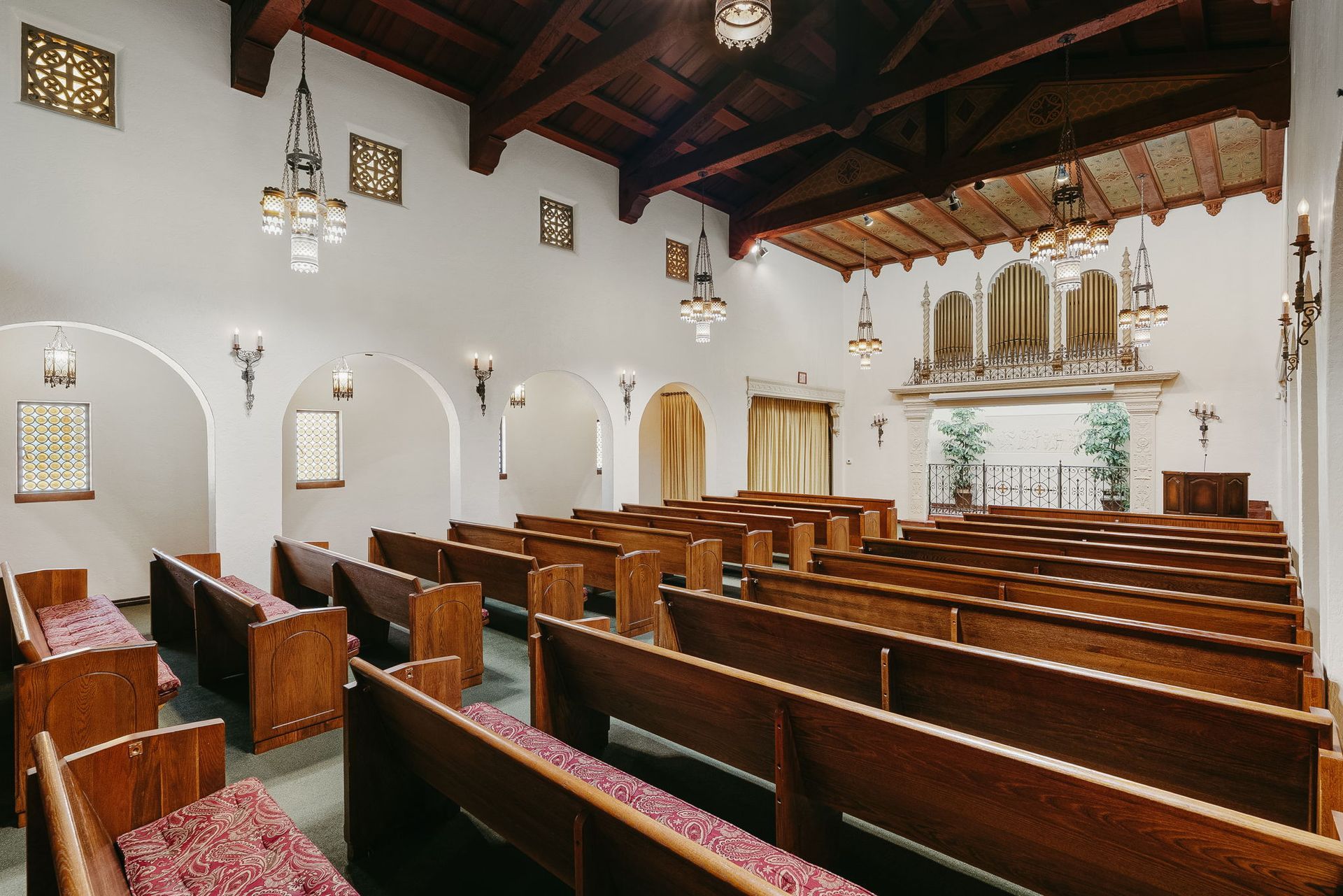 Empty church interior with wooden pews, white walls, arched doorways, and hanging chandeliers