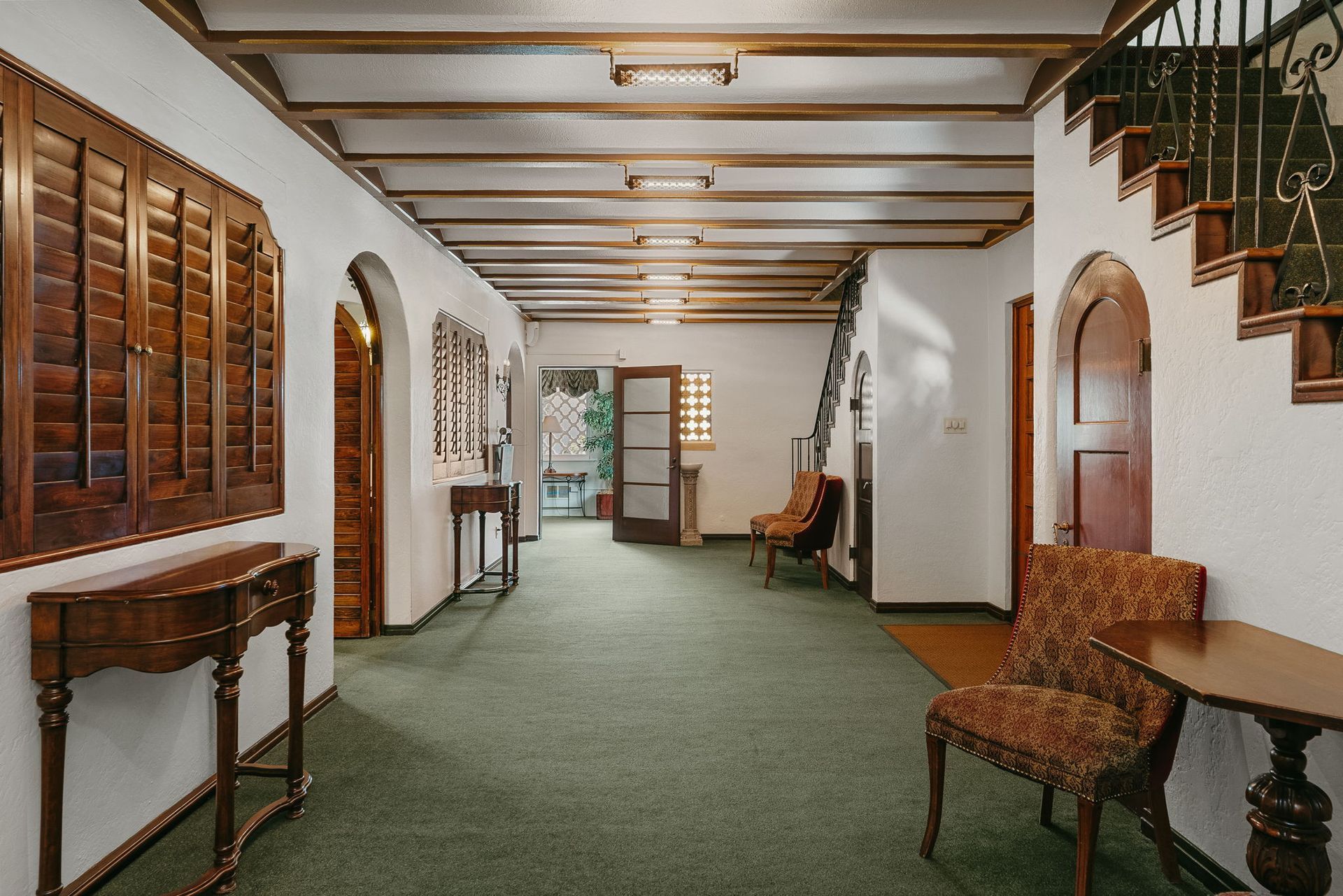 Long hallway with green carpet, white walls, wooden trim, chairs, and staircase