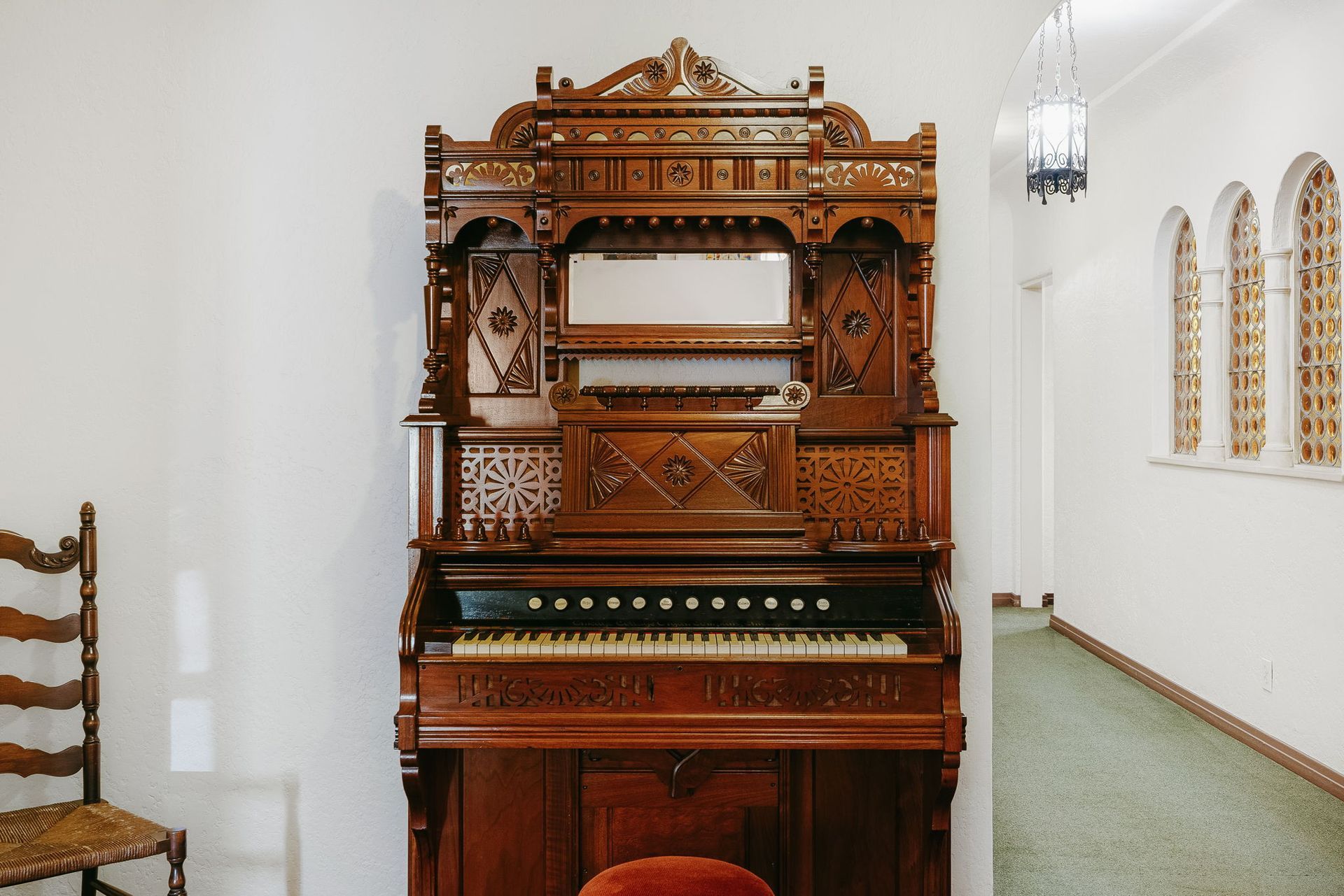 Ornate wooden piano in a white hallway, with a chair and arched windows nearby