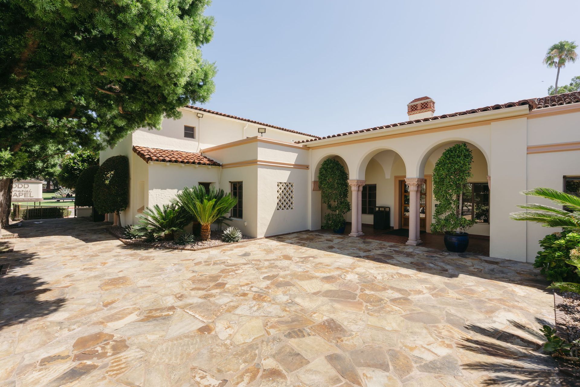 Sunlit stone courtyard outside a white stucco house with arched porch and potted plants