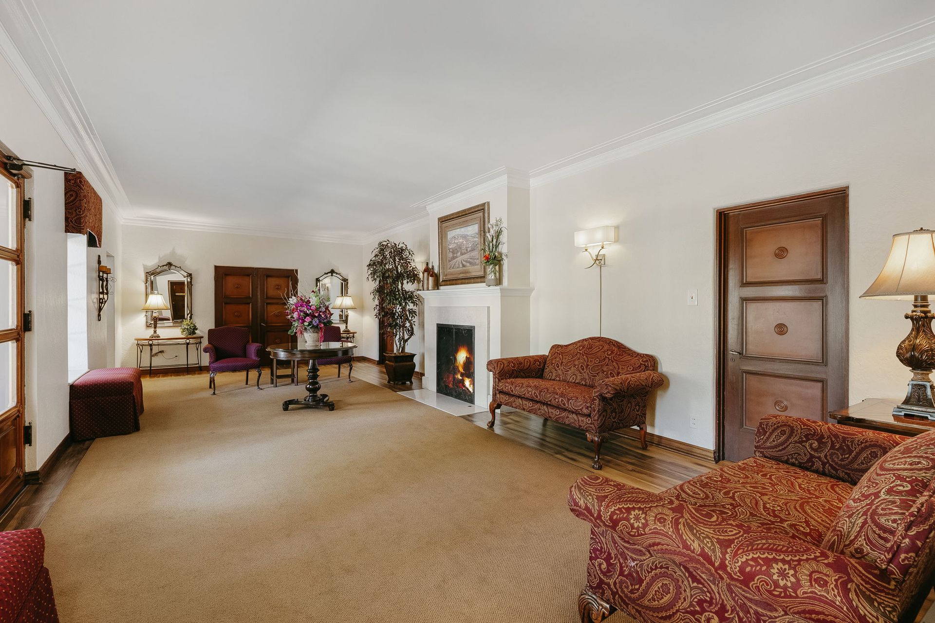 Spacious living room with beige carpet, fireplace, wooden door, and patterned armchairs.