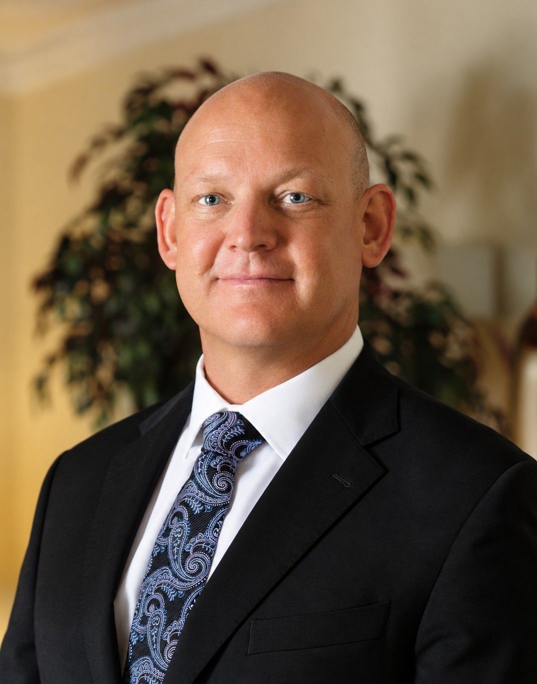 Portrait of a bald man in a black suit and blue patterned tie, standing indoors with a slight smile