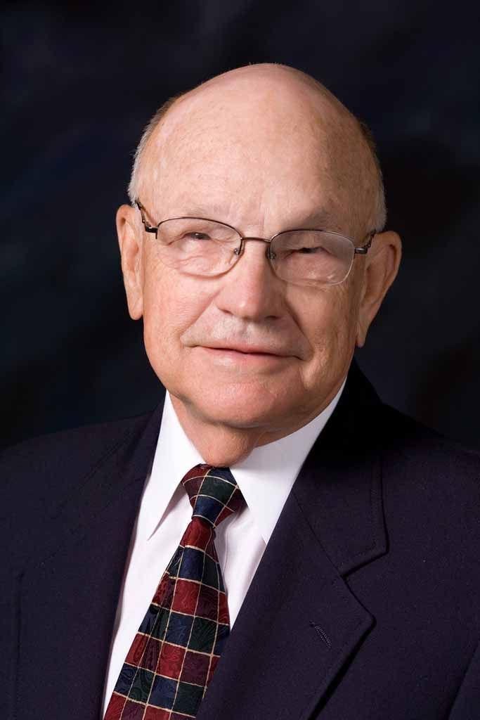 Portrait of a bald man in glasses and a suit, wearing a patterned tie against a dark background