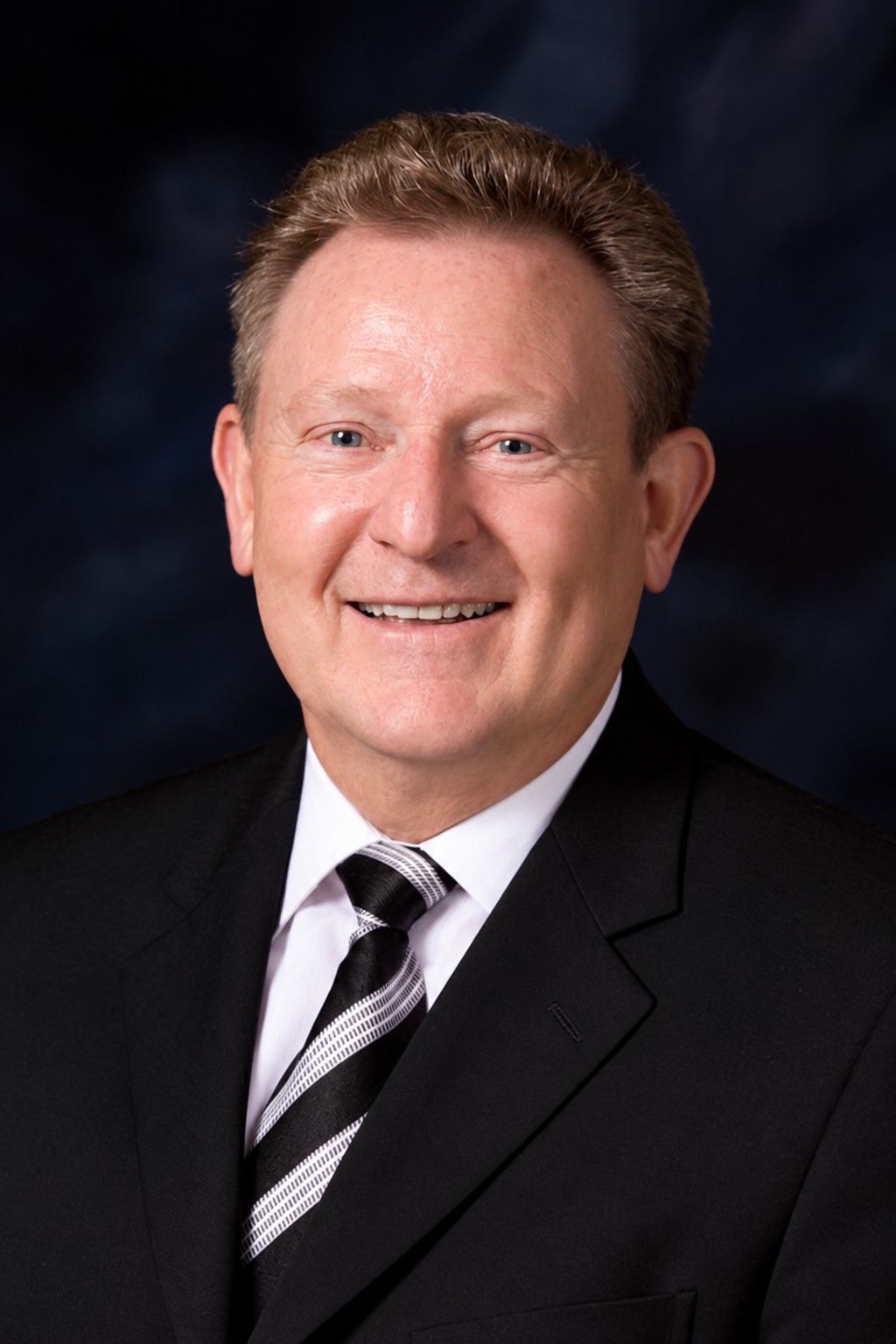 Studio portrait of a smiling man in a black suit and striped tie against a dark background