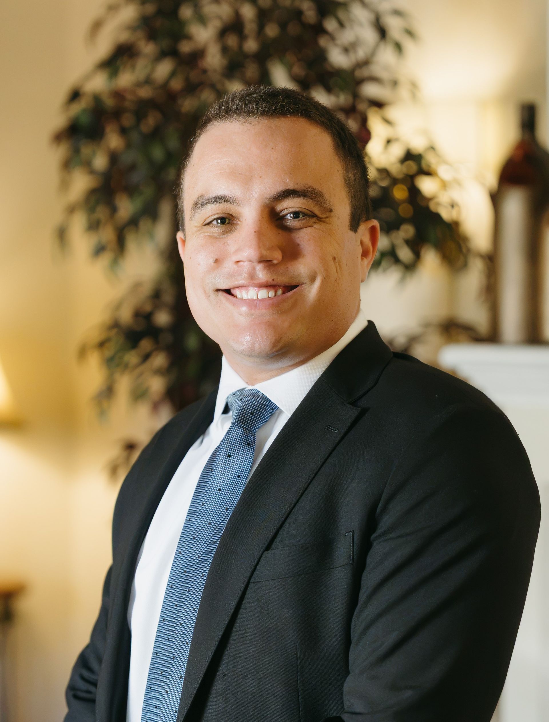 Smiling person in a dark suit and blue tie posing indoors with a blurred plant in the background