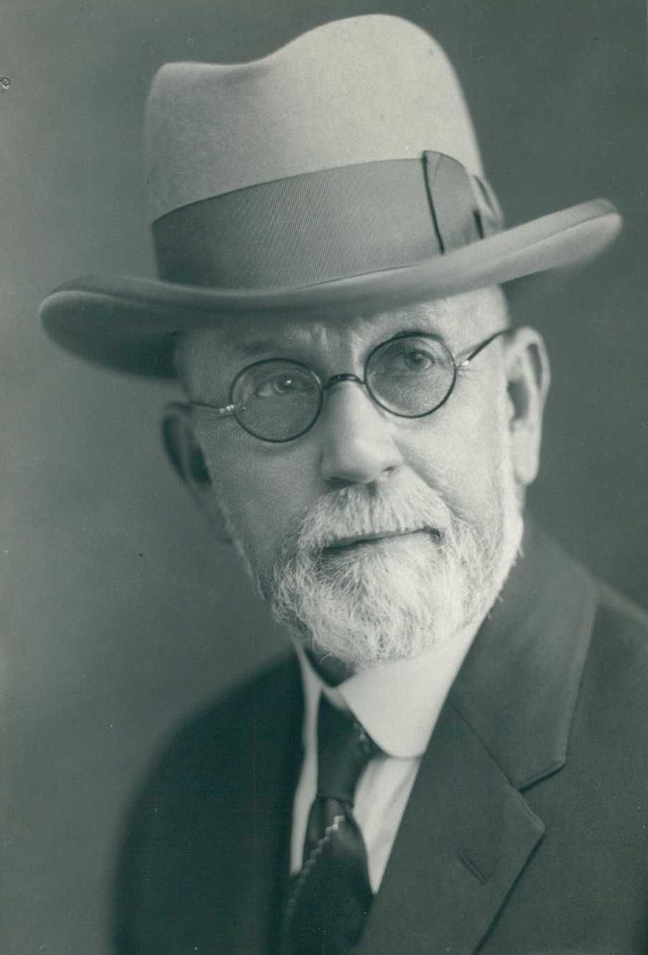 Studio portrait of a man in a hat, round glasses, and suit, facing forward with a neutral expression