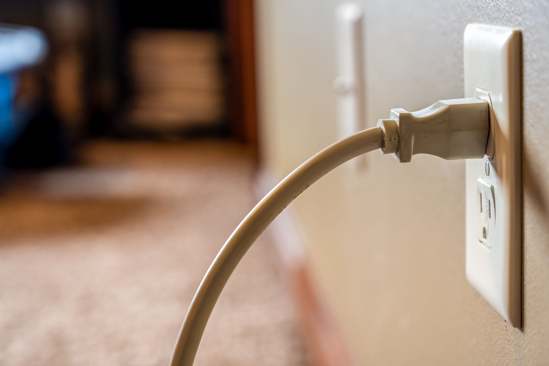 A beige power cord plugged into a white wall outlet, with a blurry room in the background.