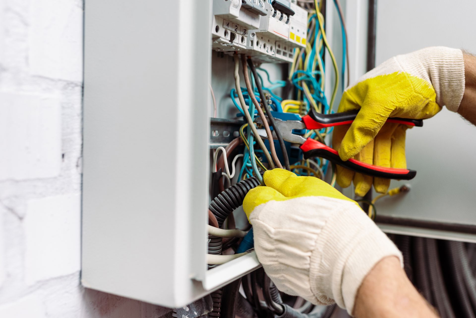 Electrician in yellow gloves wiring an open electrical panel with colored cables