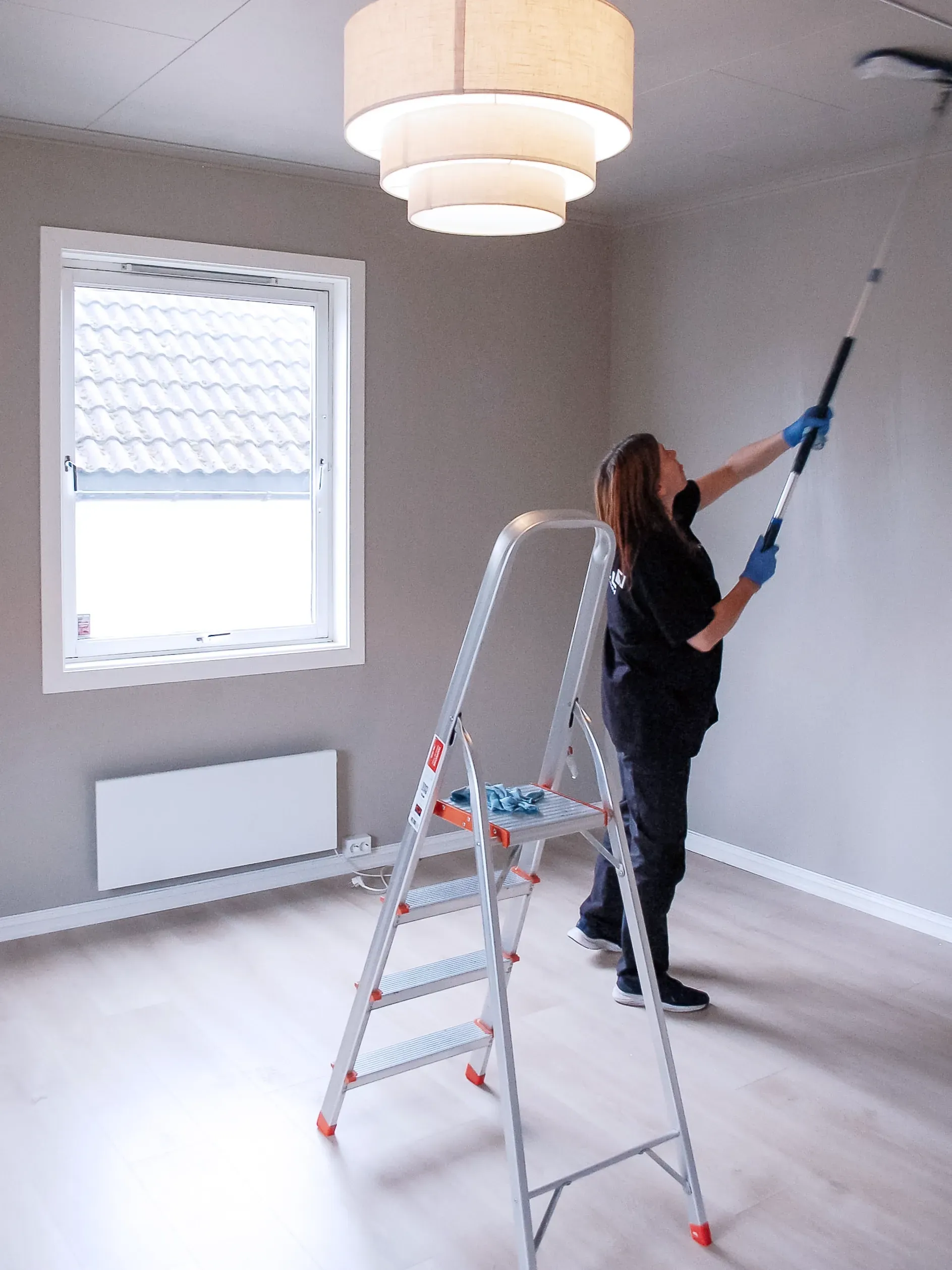 Kvinne maler en vegg med en stige i et rom med lyst gulv og en stor taklampe.